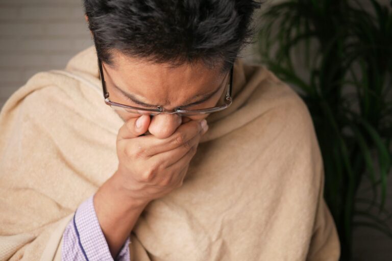 Người đàn ông đeo kính đang ho hoặc hắt hơi che miệng bằng hai tay trong khi khoác chăn