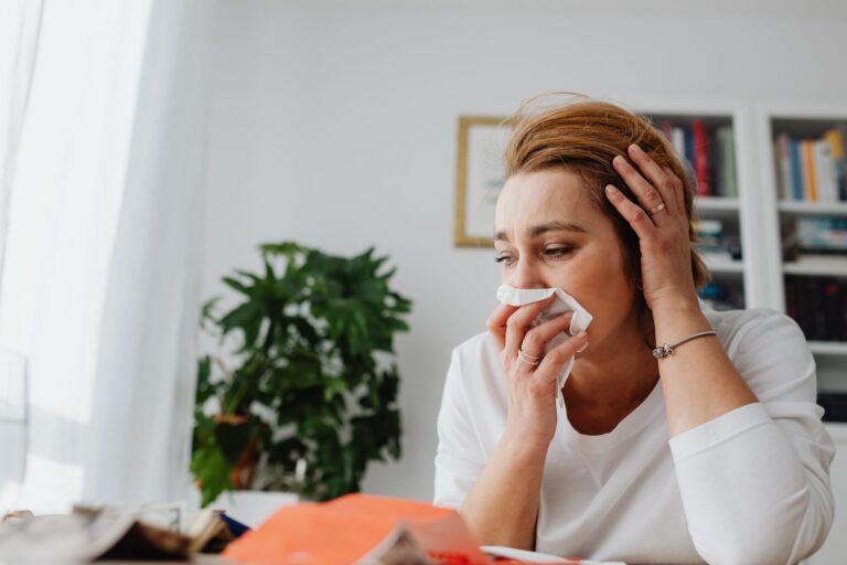 Người phụ nữ đang xì mũi bằng khăn giấy và đặt tay lên đầu vì cảm cúm