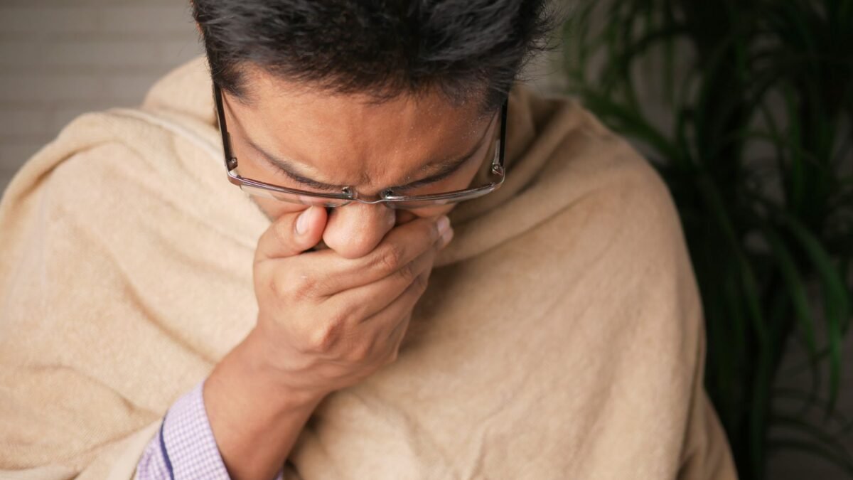 Người đàn ông đeo kính đang ho hoặc hắt hơi che miệng bằng hai tay trong khi khoác chăn